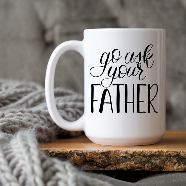 15oz white ceramic mug with hand lettered illustrated design that says go ask your father shown sitting on a wood tray with a grey knit blanket