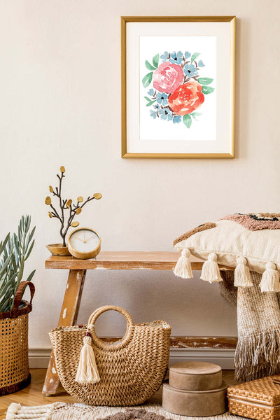 Watercolor wall art print with coral and rust colored roses, little muted blue accent flowers and green leaves shown hanging on a wall in a gold frame with white matte in a living room with a bench, pillow and neutral colored home accessories.