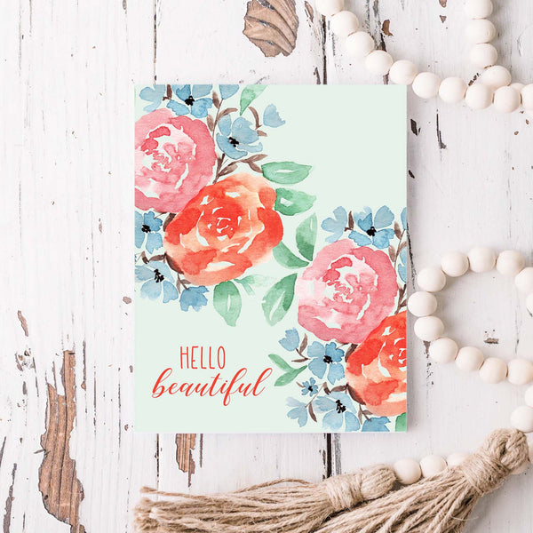 hello beautiful floral watercolor greeting card with coral, pink, blue flowers and green leaves with white A2 envelope shown on a rustic white wood table with white wood bead garland