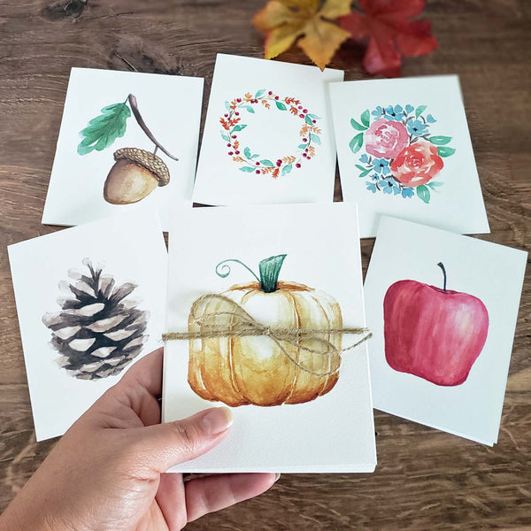 Set of 6 Watercolor painted notecards in fall autumn nature designs including an acorn with leaf, orange pumpkin, fall foliage wreath, red apple, fall floral bouquet and pinecone shown with a hand holding a set and cards in the background