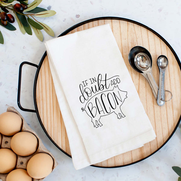 White floursack kitchen towel with black hand lettered illustrated design that says If in doubt add bacon with a outline of a pig shown folded on a serving tray with a set of measuring spoons and fresh eggs