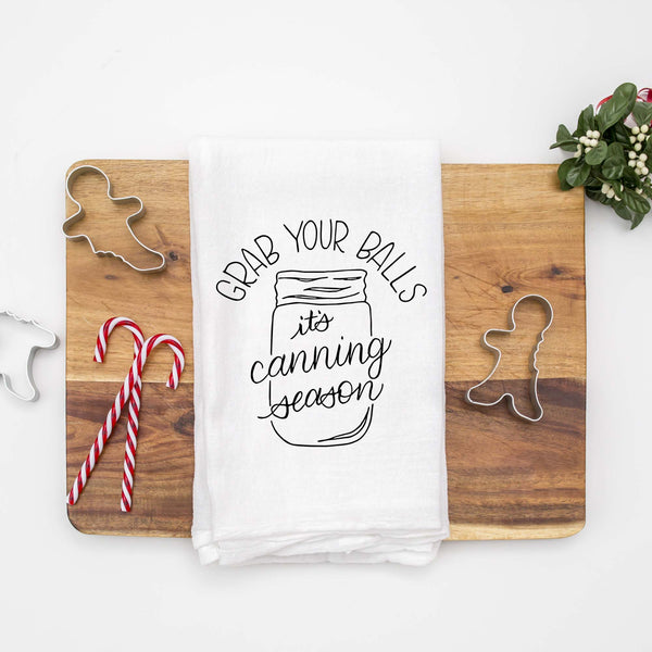 White floursack kitchen towel with black hand lettered illustrated design that says Grab your balls it's canning season with a mason jar doodle shown folded on a wooden cutting board with Christmas cookie cutters and candy canes