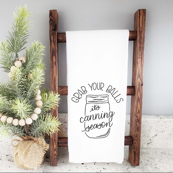 White floursack kitchen towel with black hand lettered illustrated design that says Grab your balls it's canning season with a mason jar doodle shown folded and hanging from a wooden display ladder and a mini Christmas tree