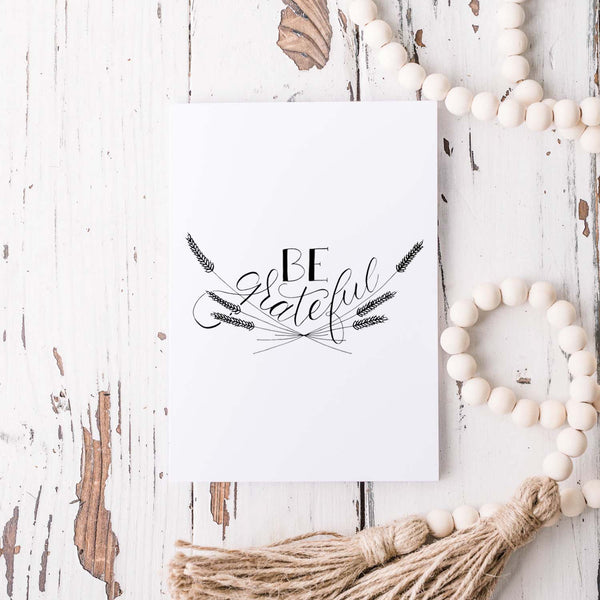 Be Grateful A2 Greeting Card, black hand lettered design with wheat illustrations on a folded white card shown on a white rustic table with a white wooden beaded garland