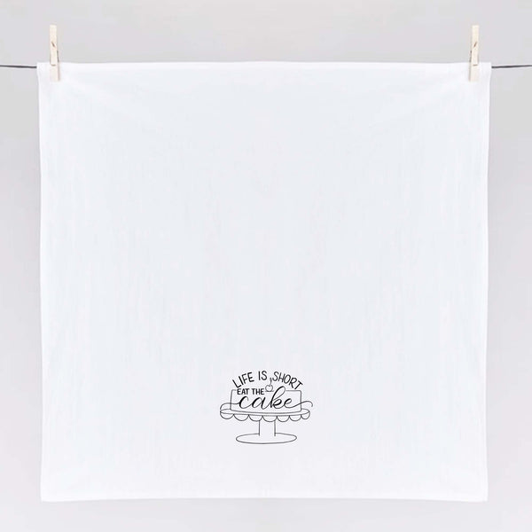 White floursack towel with black hand lettered illustrated design that says Life is short eat the cake with a cake on a stand doodle shown unfolded and hanging from clothes pins