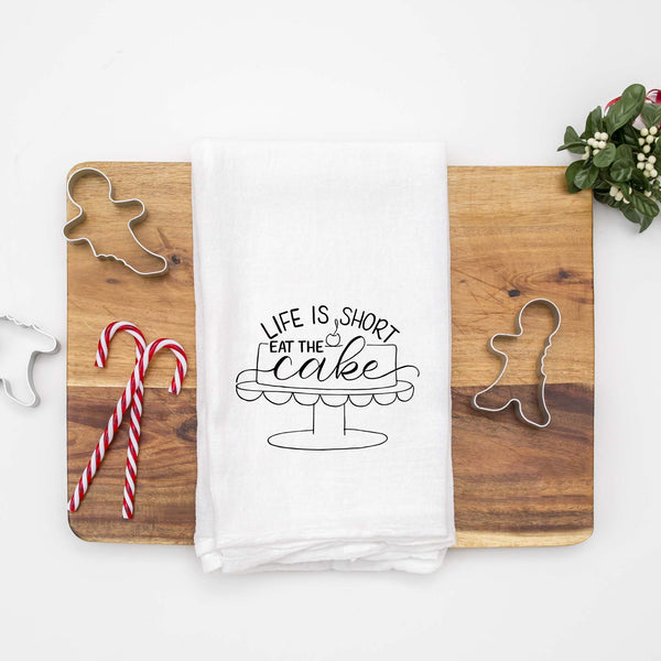 White floursack towel with black hand lettered illustrated design that says Life is short eat the cake with a cake on a stand doodle shown folded on a wood cutting board with Christmas cookie cutters and candy canes