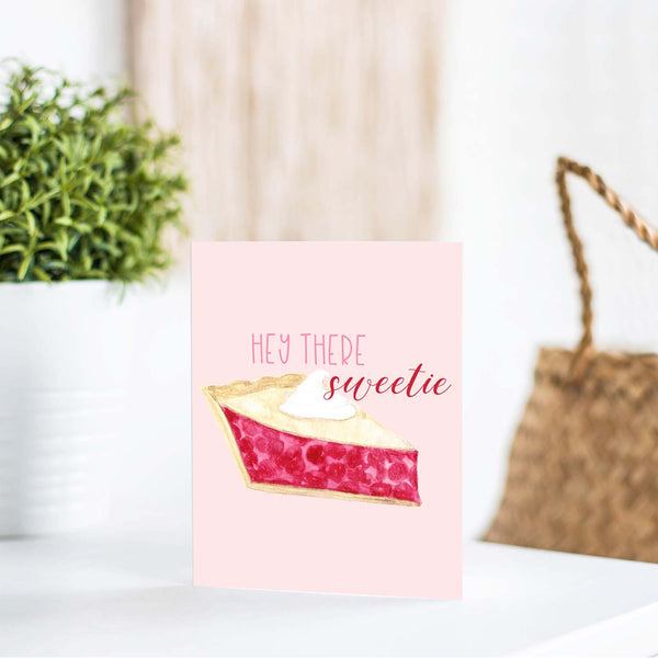 hey there sweetie watercolor greeting card with a watercolor slice of cherry pie topped with a dollop of whipped cream greeting card with white A2 envelope shown standing on a white table with a plant and handbag