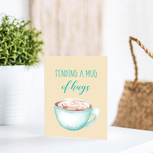 blue watercolor coffee mug filled with coffee friendship card that says sending a mug of hugs with a white A2 envelope shown standing on a white table with a plant and a handbag