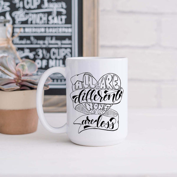 15oz white ceramic mug with hand lettered illustrated design that says all of us are different none of us are less shown in a kitchen with a plant and decorative sign