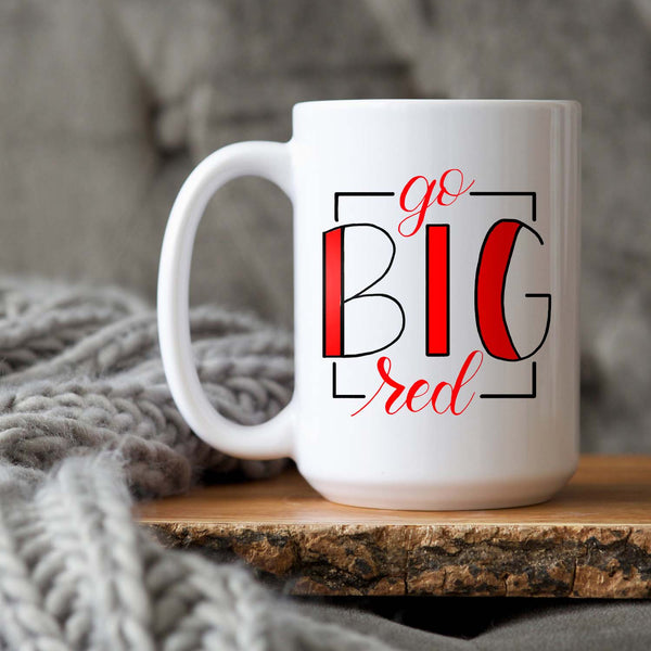 15oz white ceramic mug with hand lettered illustrated design that says Go Big Red sitting on a wood tray and a grey knit blanket