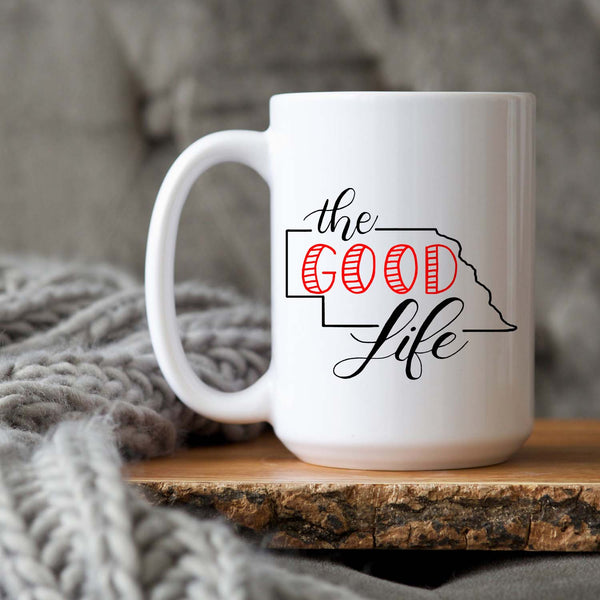 15oz white ceramic mug with hand lettered illustrated design that says The Good Life with the outline of the state of Nebraska shown sitting on wood tray and a grey knit blanket
