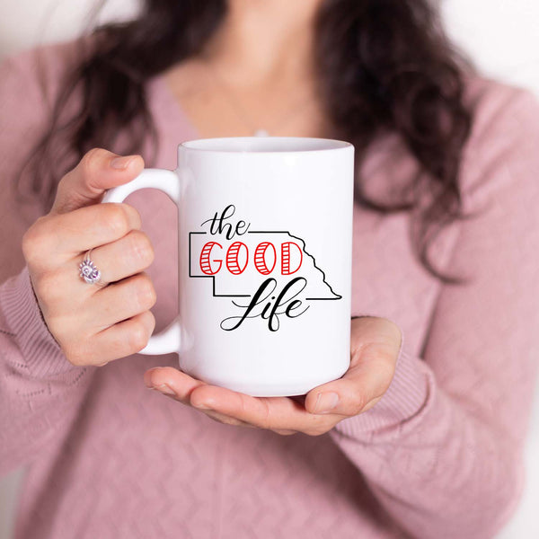 15oz white ceramic mug with hand lettered illustrated design that says The Good Life with the outline of the state of Nebraska shown with a woman holding the mug