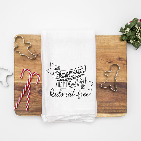 White floursack kitchen towel with black hand lettered illustration that says grandma's kitchen kids eat free shown folded on a wood cutting board with Christmas cookie cutters and candy canes