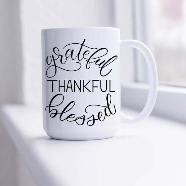15oz white ceramic mug with hand lettered illustrated design that says Grateful Thankful Blessed shown sitting in a sunny window