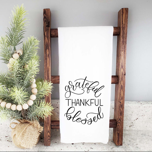 White floursack towel with black hand illustrated design that says grateful thankful blessed shown folded and hanging from wooden display ladder with a mini Christmas tree