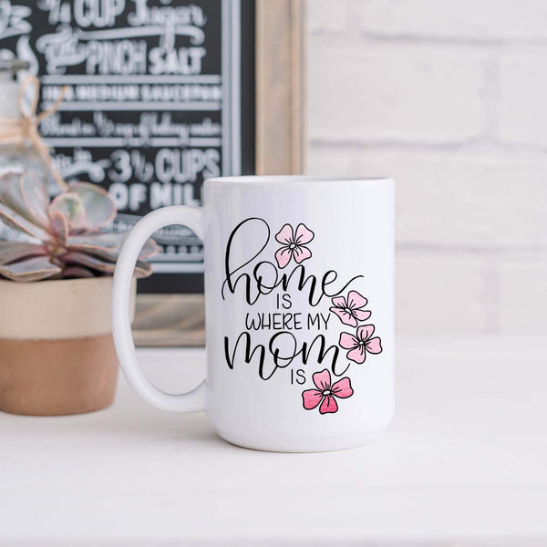 15oz white ceramic mug with hand lettered illustrated design that says home is where my mom is with pink flower illustrations shown in a kitchen