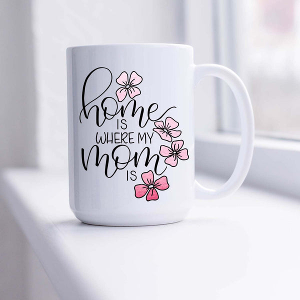 15oz white ceramic mug with hand lettered illustrated design that says home is where my mom is with pink flower illustrations shown sitting in a sunny window