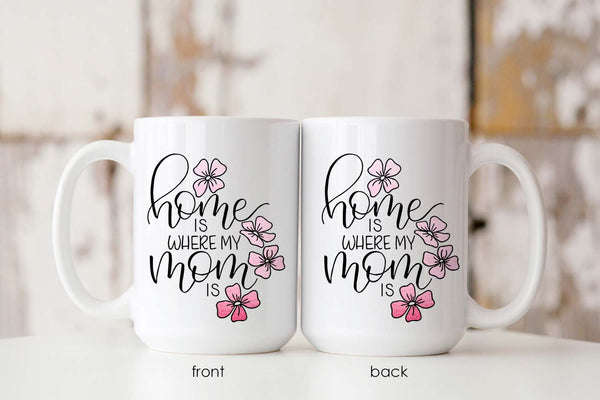 15oz white ceramic mug with hand lettered illustrated design that says home is where my mom is with pink flower illustrations showing both front and back of the mug