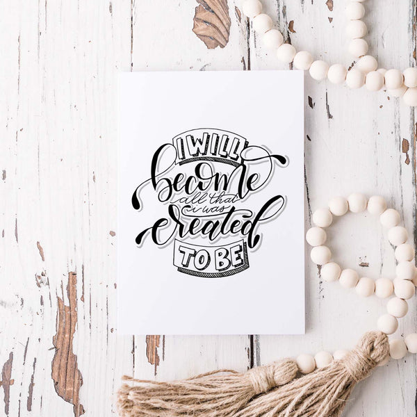 I will become all that I was created to be black and white hand lettered greeting card on a white folded card with A2 envelope shown on a white rustic table with a white wooden bead garland