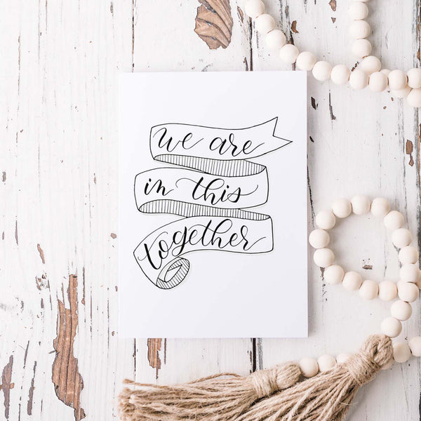 we are in this together hand lettered and illustrated black and white greeting card on a white folded card with A2 envelope shown on a white rustic table with a white wooden bead garland