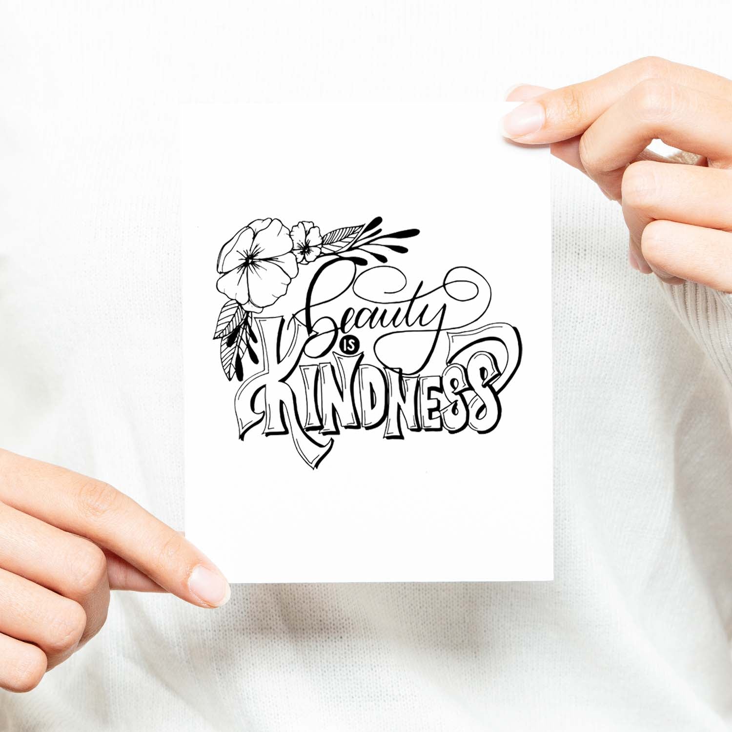 Beauty is kindness hand lettered and illustrated black and white greeting card on a folded white card with an A2 envelope shown with a woman in a white sweater holding card