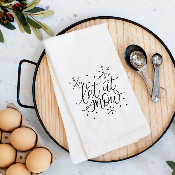White Floursack Kitchen Towel with black hand lettered illustration that says let it snow with snowflake doodles shown folded on a serving tray with a set of measuring spoons and fresh eggs in a modern kitchen