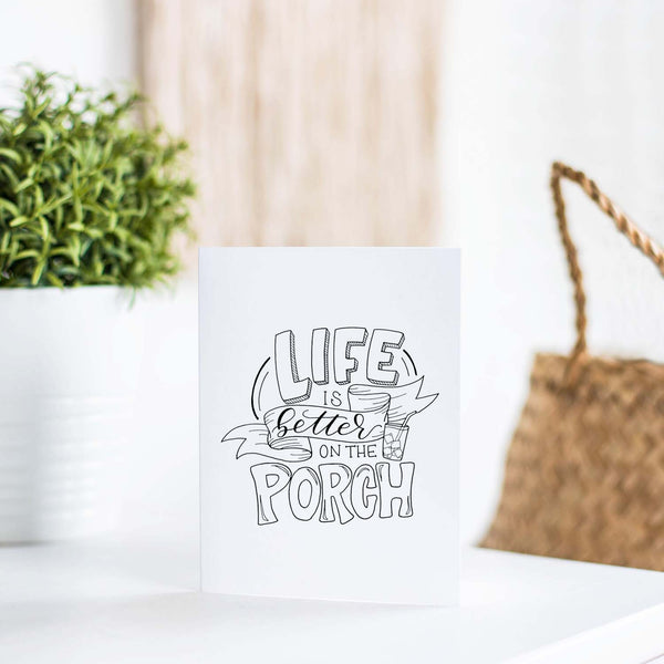 life is better on the porch hand lettered and illustrated black and white greeting card with A2 envelope shown on a white table with plant and handbag