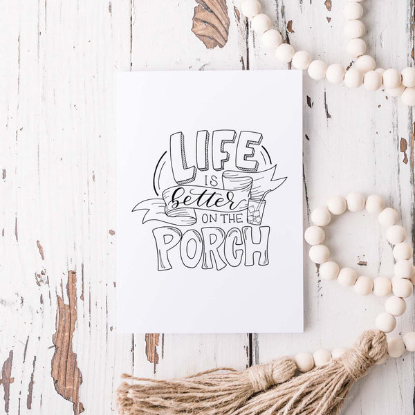 life is better on the porch hand lettered and illustrated black and white greeting card with A2 envelope shown on a white rustic table with a white wooden bead garland