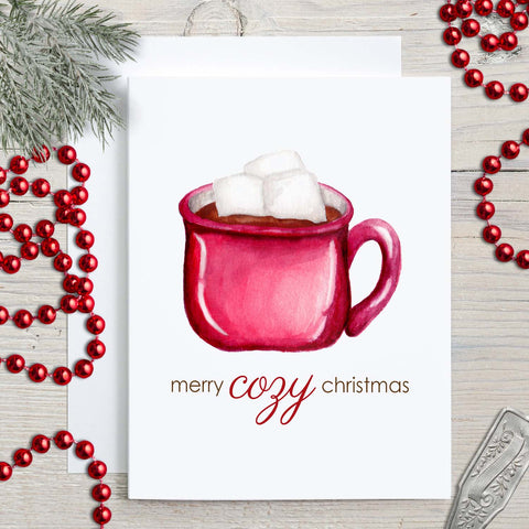 Watercolor Christmas greeting card with a watercolor painted red mug full of hot  cocoa and heaping with marshmallows that says merry cozy christmas shown with holiday decorations