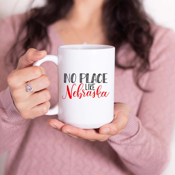 15oz white ceramic mug with hand lettered illustrated design that says No Place Like Nebraska shown with a woman holding the mug