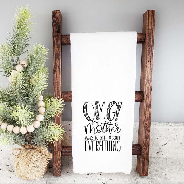 White floursack kitchen towel with black hand lettered illustrated design that says OMG! My mother was right about everything shown folded and hanging from a wooden display ladder and a mini Christmas Tree