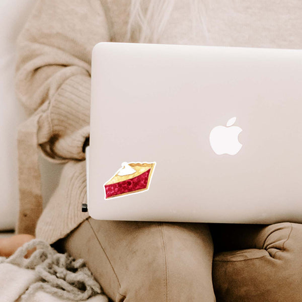 3" vinyl sticker of a watercolor painted slice of cherry pie with whipped cream on top shown adhered to a MacBook cover sitting open on a woman's lap