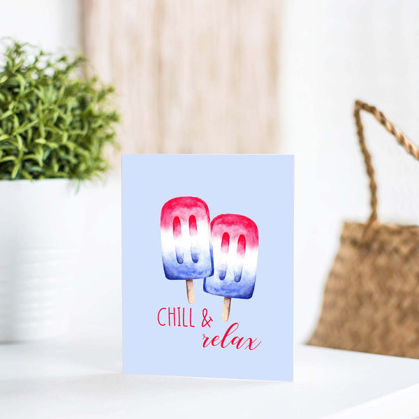 watercolor red white and blue summertime popsicles on a friendship greeting card that says chill & relax with a white A2 envelope shown standing up on a white table with a plant and handbag