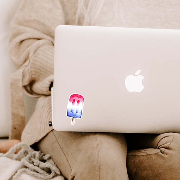 3" vinyl sticker of a watercolor painted red, white and blue patriotic popsicle shown adhered to a MacBook cover sitting open on a woman's lap