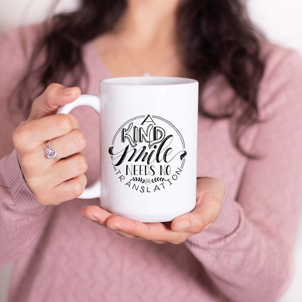 15oz white ceramic mug with hand lettered illustrated design that says a kind smi/le needs no translation shown with a woman holding the mug