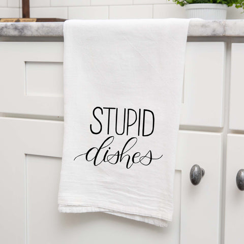 White floursack towel with black hand lettered illustrated design that says Stupid dishes shown folded and hanging from a countertop in a modern kitchen