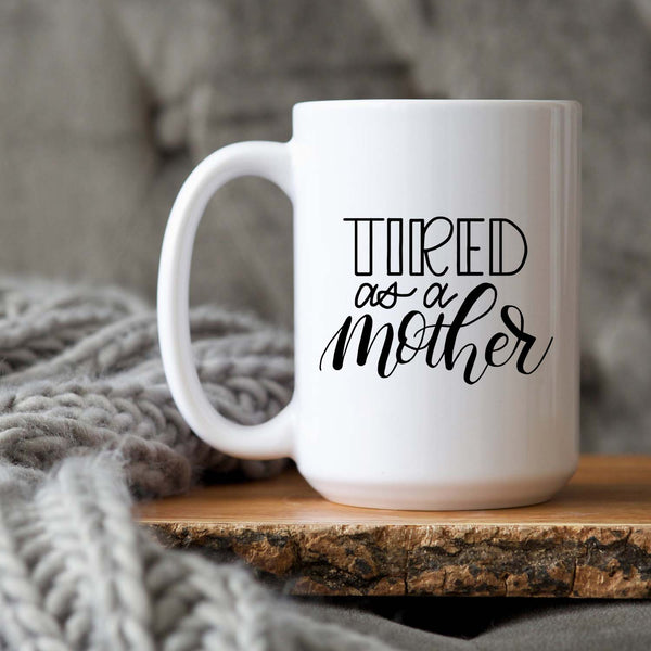 15oz white ceramic mug with hand lettered illustrated design that says tired as a mother shown sitting  on a wood tray with a grey knit blanket