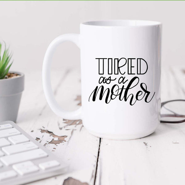 15oz white ceramic mug with hand lettered illustrated design that says tired as a mother shown sitting on a white office desk