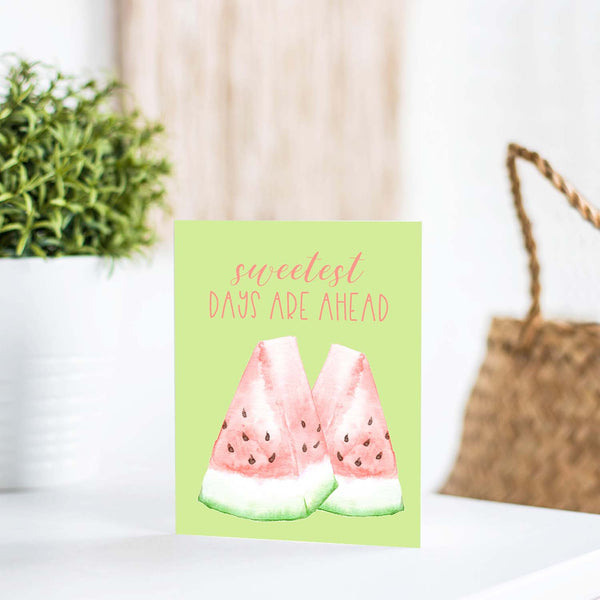 watercolor pink and green watermelon slices on a greeting card that says sweetest days are ahead with a white A2 envelope shown standing on a white table with a plant and handbag