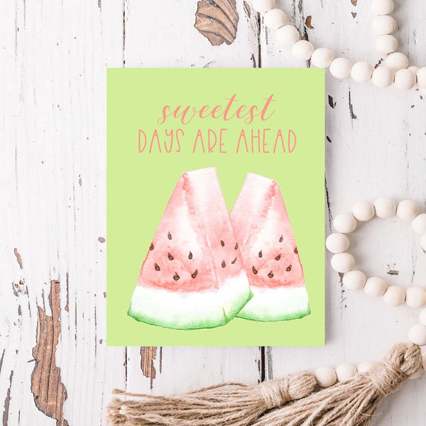 watercolor pink and green watermelon slices on a greeting card that says sweetest days are ahead with a white A2 envelope shown laying on a rustic white wooden table with white wood bead garland