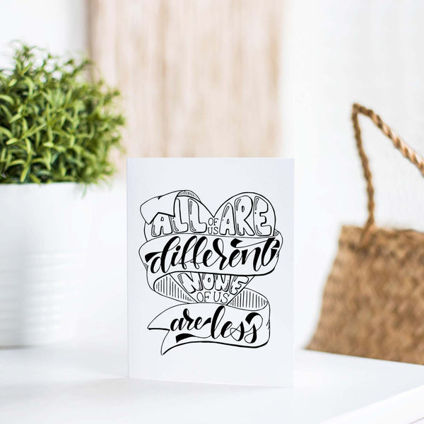 all of us are different none of us are less hand lettered and illustrated black and white on a white folded greeting card with A2 envelope shown on a white  table with a plant and handbag