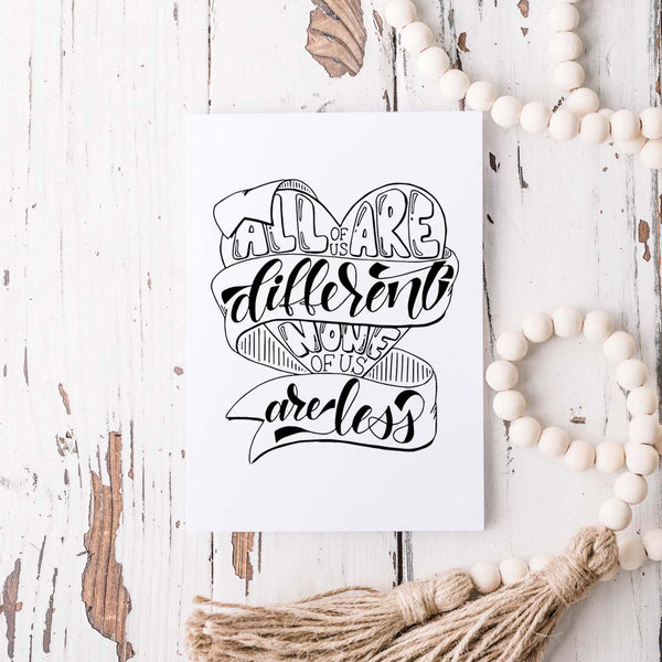 all of us are different none of us are less hand lettered and illustrated black and white on a white folded greeting card with A2 envelope shown on a white rustic table with a wooden bead garland
