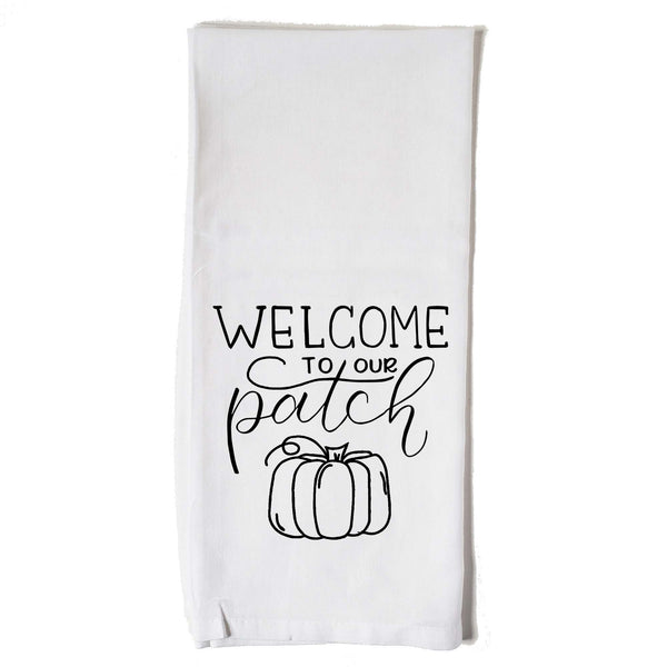 White floursack towel with black hand lettered illustrated design that says Welcome to our patch with pumpkin doodle
