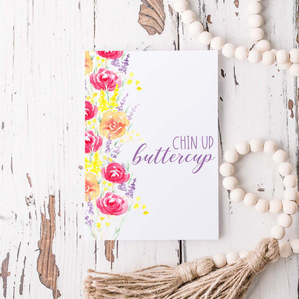 summery watercolor floral bouquets on an encouragement greeting card that says chin up buttercup with a white A2 envelope shown laying on a rustic white wooden table with  white wood bead garland