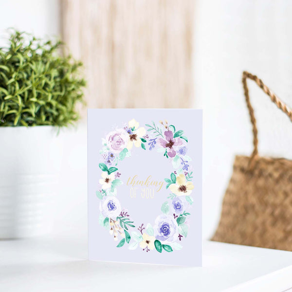 watercolor wild flower wreath on a friendship greeting card that says thinking of you in the center with a white A2 envelope shown standing on a white table with a plant and handbag