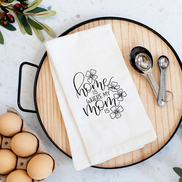 White floursack towel with black hand lettered illustrated design that says home is where my mom is with flower doodles shown folded on a serving tray with a set of measuring spoons and fresh eggs