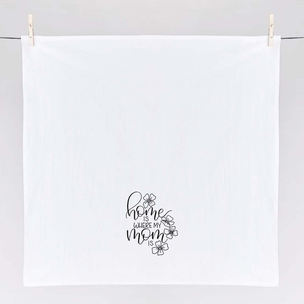 White floursack towel with black hand lettered illustrated design that says home is where my mom is with flower doodles shown unfolded and hanging from clothes pins