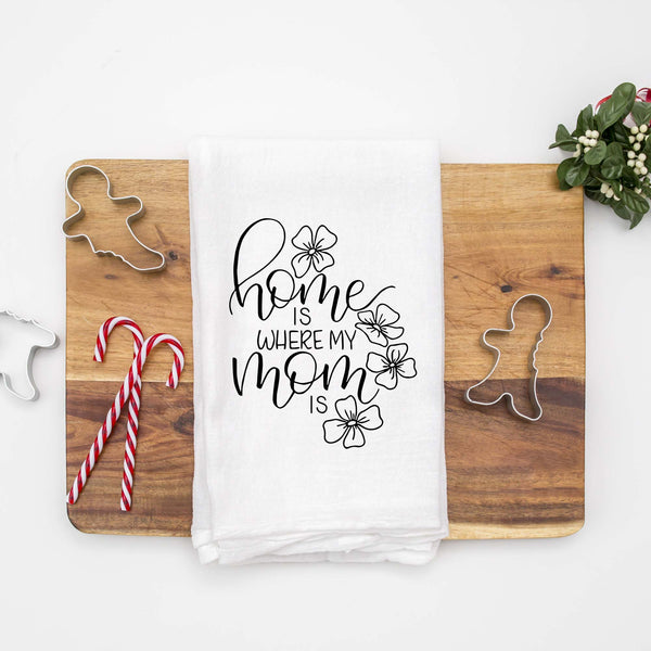 White floursack towel with black hand lettered illustrated design that says home is where my mom is with flower doodles shown folded on a wood cutting board with Christmas cookie cutters and candy canes
