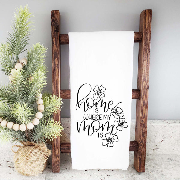 White floursack towel with black hand lettered illustrated design that says home is where my mom is with flower doodles shown folded on a wooden display ladder with a mini Christmas Tree