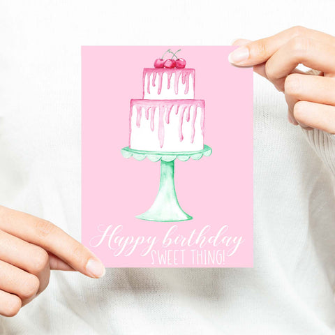 Happy birthday sweet thing watercolor birthday cake with pink frosting cherries and seafoam green cake stand birthday card shown with a woman in a white sweater holding card